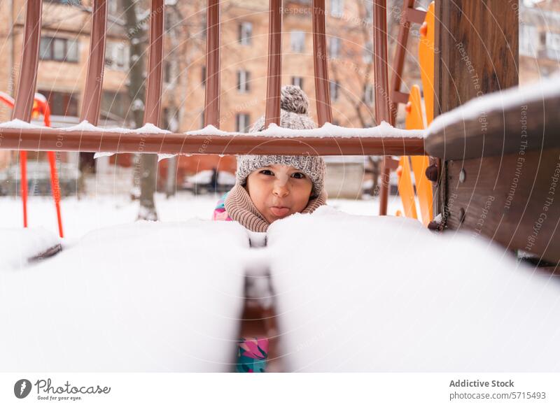 Curious child peeking through snow-covered playground equipment in winter attire Child curious outdoor cold scarf knitted hat enjoyment rosy cheek frosty
