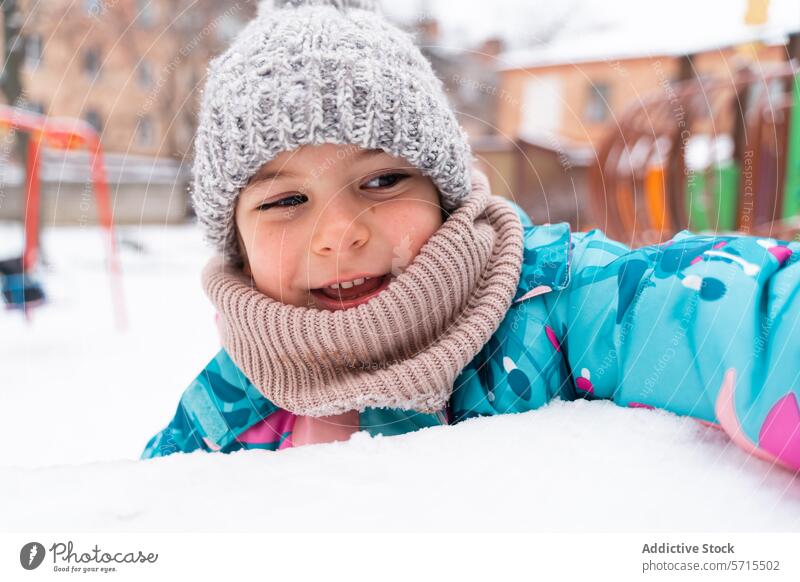 Smiling child lying in the snow, wearing a knitted hat and colorful jacket, with playground in view Child smile winter outdoor cold happy fun scarf joy knitwear