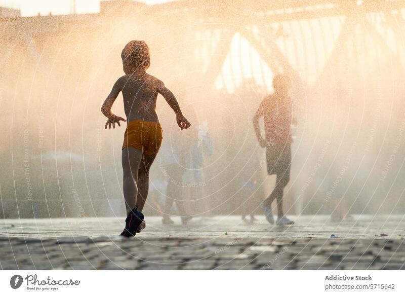 Anonymous silhouette of a child runs through the mist of a city fountain as the sun descends, casting a warm, golden glow over the urban landscape spray dusk