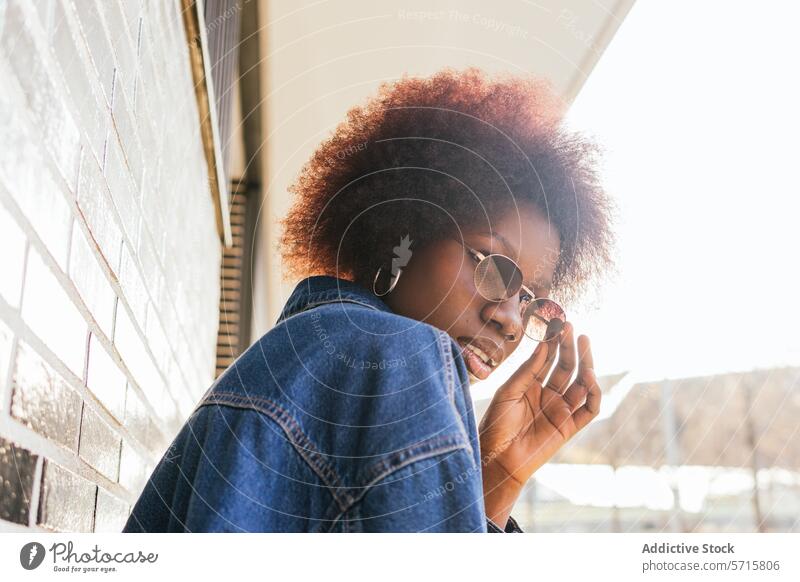 A contemplative young woman adjusts her round sunglasses, her afro haloed by the sun against a brick wall backdrop denim jacket reflective mood black ethnic