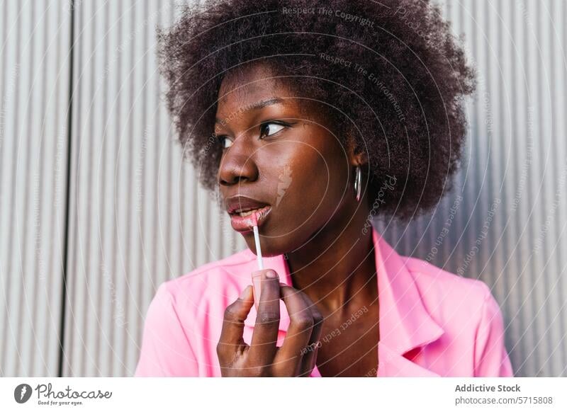 A fashionable black woman applies lip gloss with precision, her expression focused, highlighting a moment of personal style in an urban setting application pink