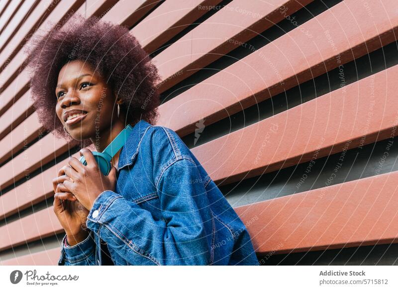 A radiant young woman smiles, headphones around her neck, set against a geometric background, embodying urban cool denim jacket African American casual style
