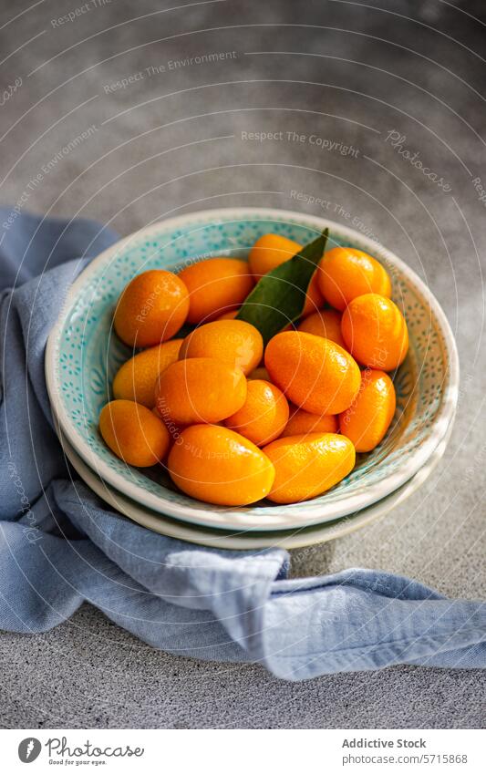 Top view of bowl of vibrant, fresh kumquats with a green leaf, placed on a textured surface with a blue napkin Kumquat fruit citrus healthy organic snack ripe