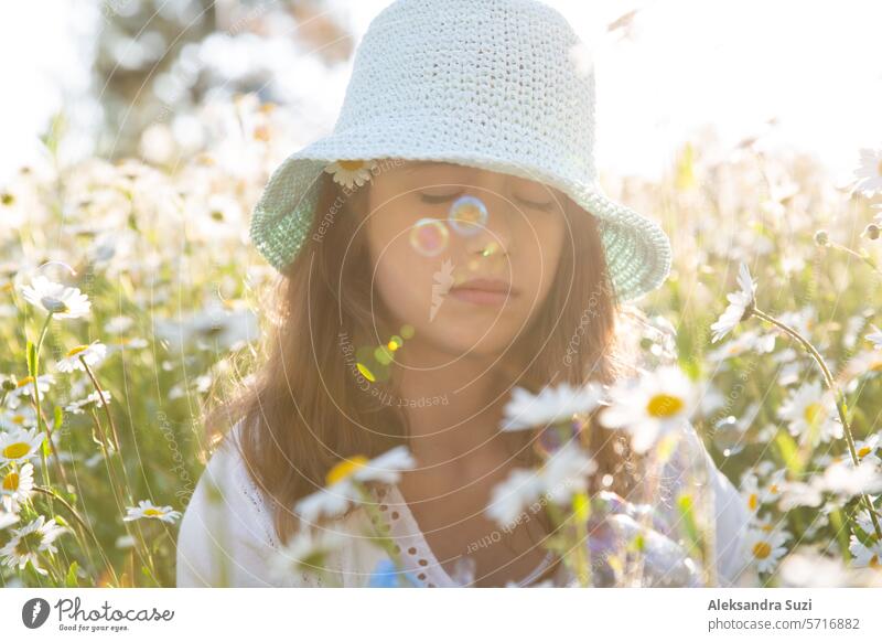 Girl sitting on a meadow covered with wild flowers. Background of field of daisy flowers in bloom. Sunny summer day, rays of light. Summer vacation concept