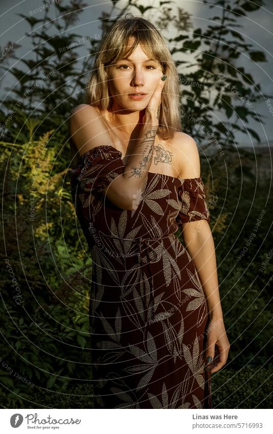 A stunning blonde girl with beautiful tattoos is basking in the warm sun rays. She wears a summer dress, creating a sensual atmosphere with a fine model-like pose. A true beauty of a pretty woman.