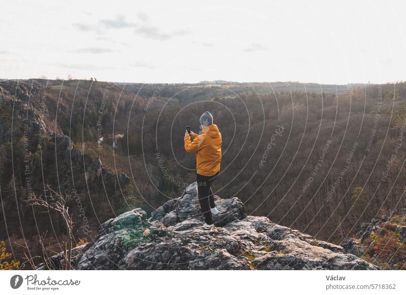 traveller in a yellow jacket standing on the edge of a cliff taking a picture of the landscape on his mobile phone at sunset. A memory of a trip. Wild Sarka Valley, Prague