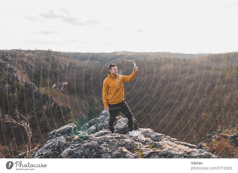 Traveller in a yellow jacket standing on the edge of a cliff taking a selfie on his mobile phone at sunset.  Divoka Sarka valley, Prague smile person