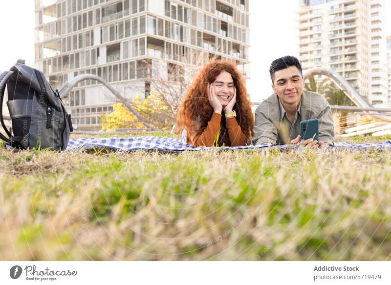 Young couple enjoying a picnic in urban park man woman smartphone city outdoor blanket grass building smiling leisure recreation happy casual relaxation