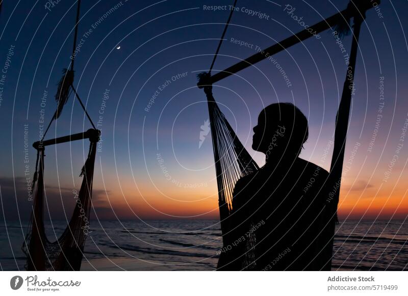 Anonymous person silhouette stands next to a hammock on the beach, with the last light of sunset and a crescent moon above twilight serenity sea dusk relaxation