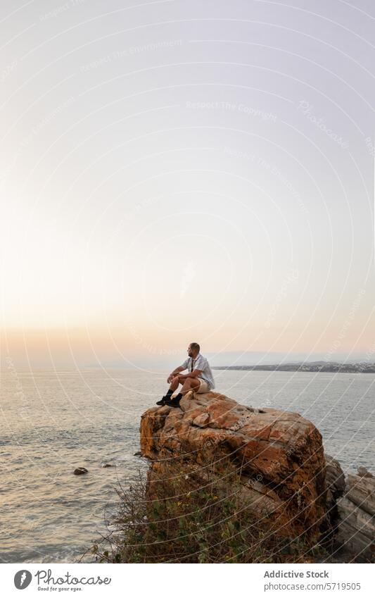 An individual sits atop a rugged cliff, gazing out at the ocean, immersed in the tranquility of a soft sunset solitude reflection sitting coast sea calm serene