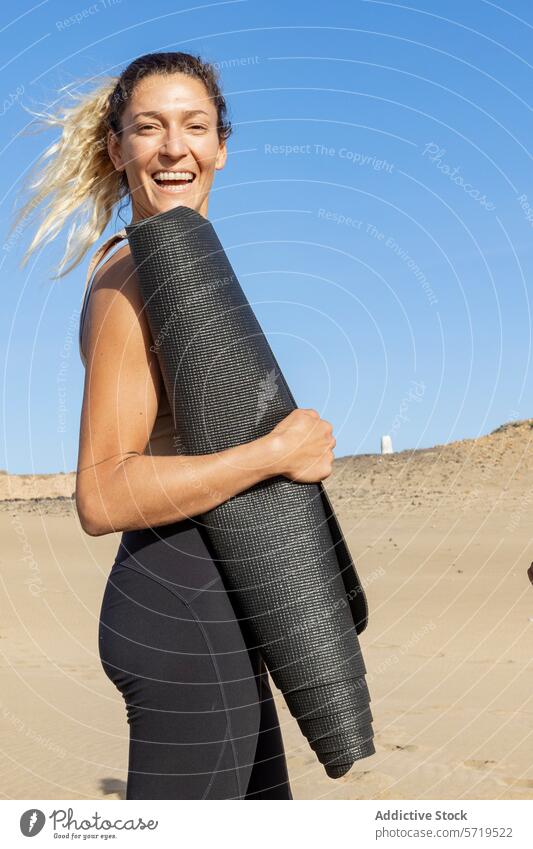 Smiling woman holding yoga mat on beach smiling healthy active fitness sunny outdoors lifestyle exercise wellness joyful active wear sport sand leisure athletic