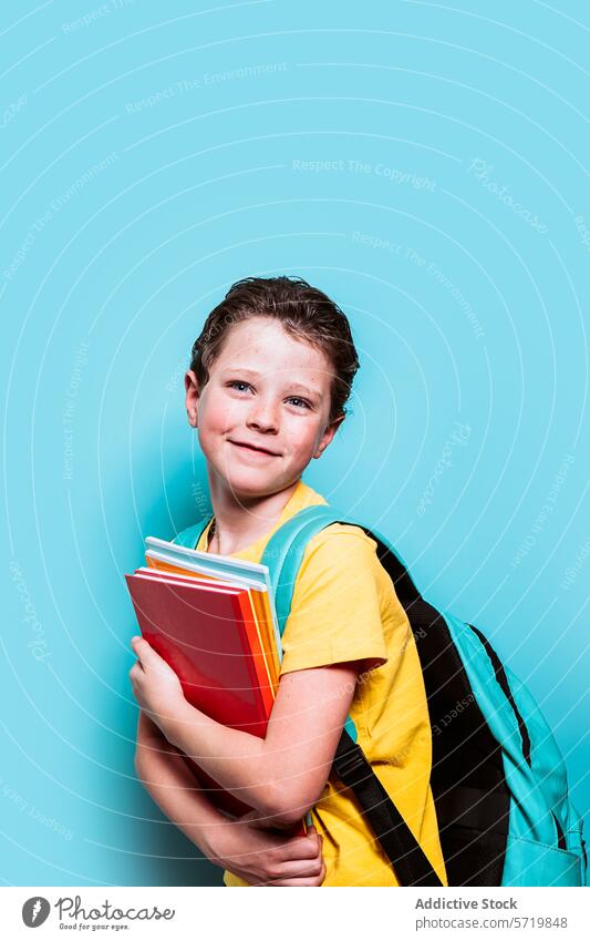 A bright-eyed boy with a sincere smile carries a stack of books and his backpack, standing before a playful blue backdrop school yellow shirt child student