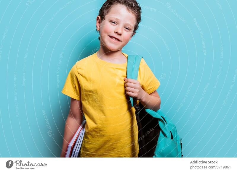 A young student in a yellow tee grins while holding textbooks and wearing a backpack, set against a light blue background boy school smile education learning
