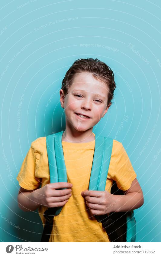 A joyful school-aged boy in a yellow shirt beams with a bright smile, holding onto his backpack straps, ready for a school day happy turquoise background child