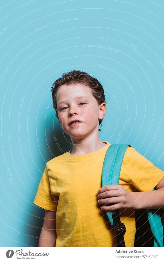A young schoolboy in a yellow shirt grips his teal backpack strap, standing before a soft blue background, looking thoughtful student child education elementary