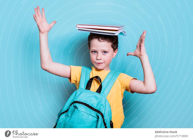 A school boy with a backpack strikes a playful pose, balancing a stack of books on his head against a light blue background balance student child education fun