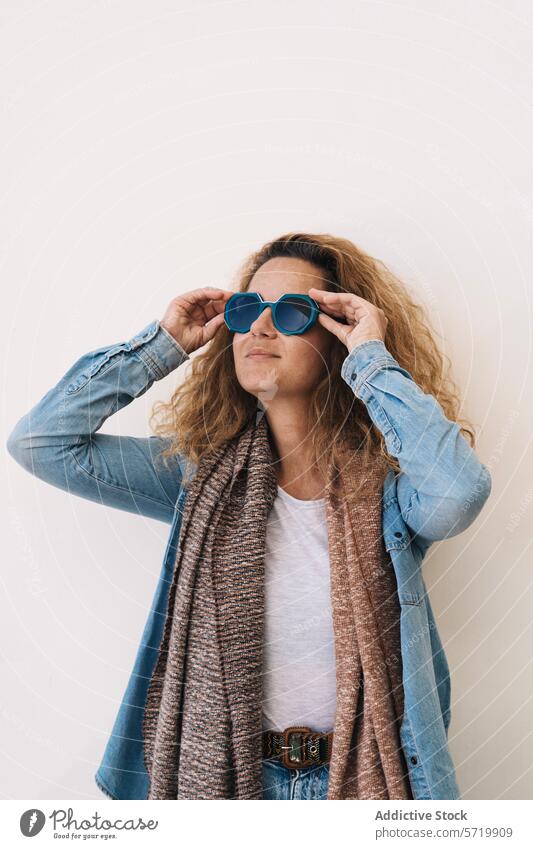 A stylish woman in a denim jacket and knitted scarf confidently adjusts her oversized blue sunglasses against a plain background style fashion chic casual