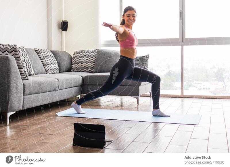 Young Woman Practicing Warrior II Pose Indoors online with tablet young adult female hispanic warrior ii pose virabhadrasana ii indoor yoga session sunlight
