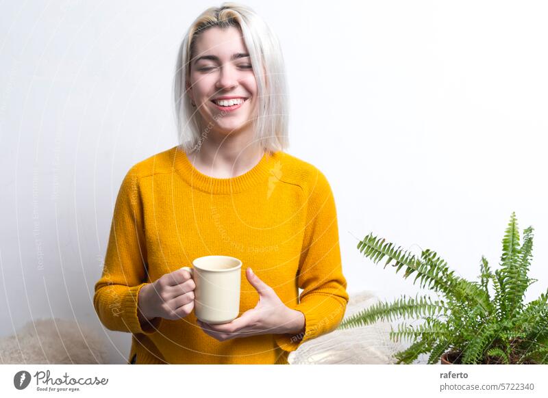 Joyful woman holding a mug with eyes closed. joyful yellow sweater coffee smile content warm happy relaxation break home indoor plant leisure cozy comfort