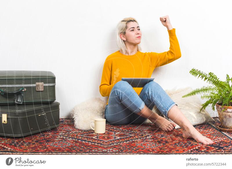 Woman with tablet making a fist of success at home. woman yellow sweater victory triumph casual relaxed denim sitting floor celebration achievement modern