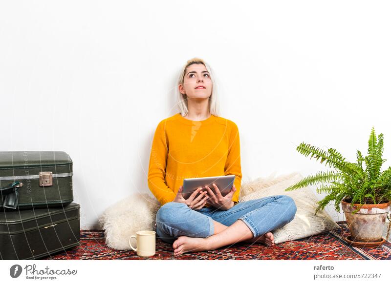 Contemplative woman with tablet looking upwards at home. contemplation inspiration yellow sweater thinking pensive curious denim jeans sitting cozy indoor