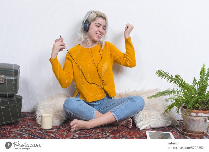 Woman with headphones dancing and holding phone. woman smartphone happy yellow sweater music joy home energy casual denim jeans sitting rug enjoyment technology