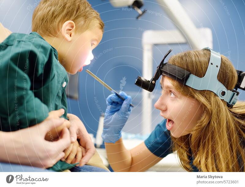 A cute toddler boy with his mother are at an appointment with a pediatric ENT. The doctor otolaryngologist is examines the throat of a little patient with inspection spatula