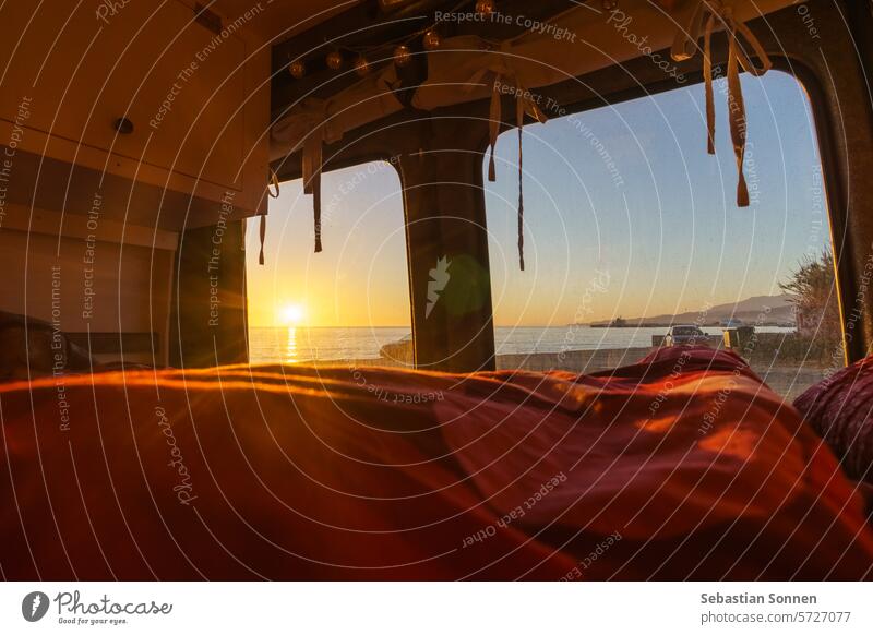 View out of a self built camper van at a beach of the mediterranean sea with colorful sunset over the water surface, Andalusia, Spain home travel light wood bed
