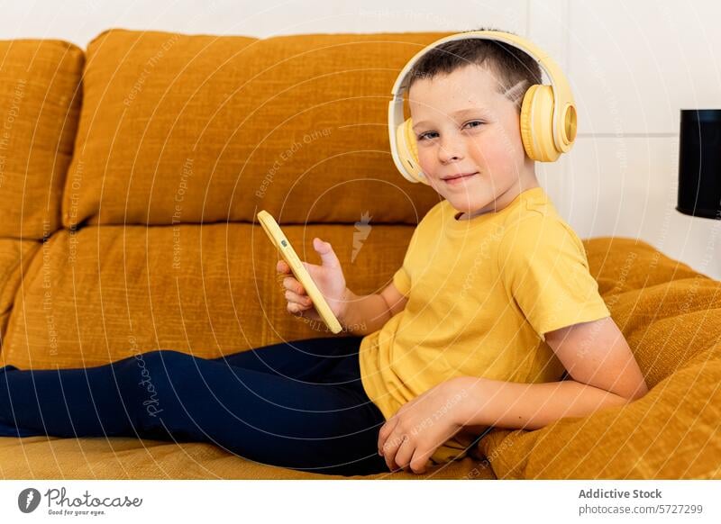 Young boy enjoys music on a tablet at home child headphones sofa leisure technology cheerful lounging mustard yellow holding device digital entertainment
