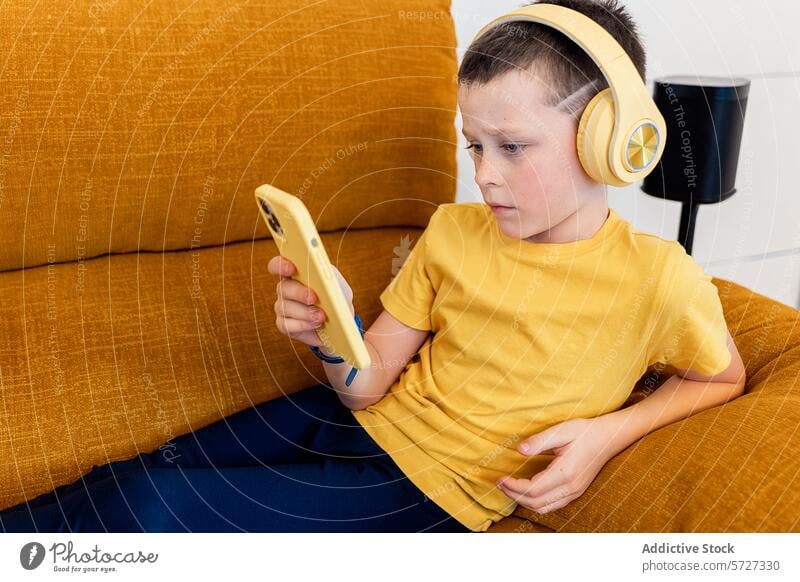 Young boy in headphones focused on smartphone at home couch young sitting technology child digital screen time yellow mustard absorbed indoor leisure activity