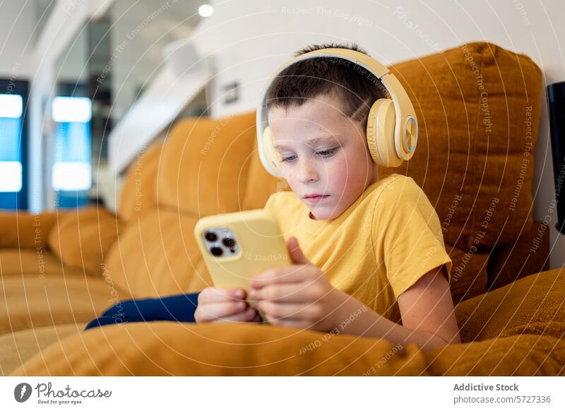 Young boy engrossed in smartphone with headphones couch mustard concentrated engaged yellow sitting technology screen digital device mobile young child kid