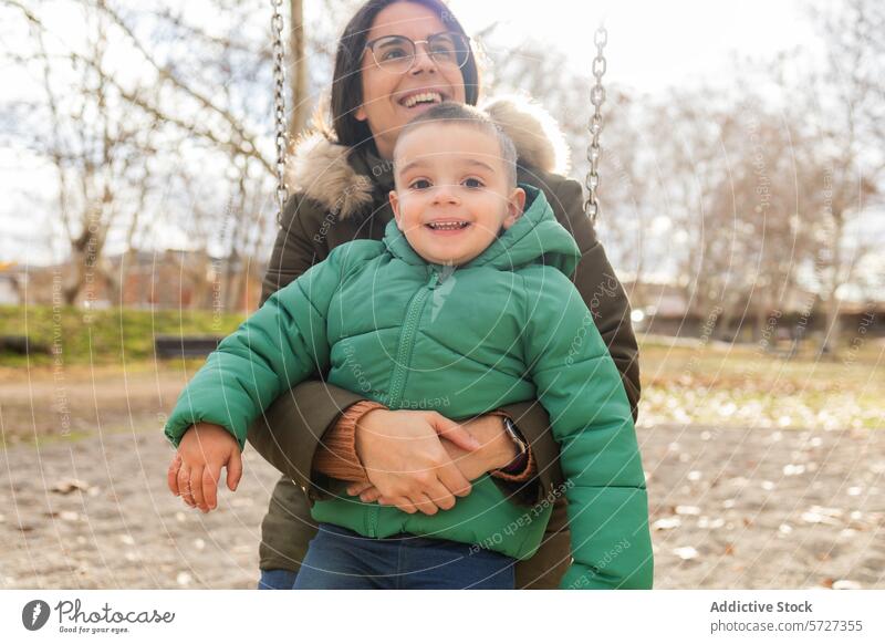 A little boy beams with happiness on a swing, securely held by his mother, both enjoying a bright day at the park outdoor fun child family smile happy play