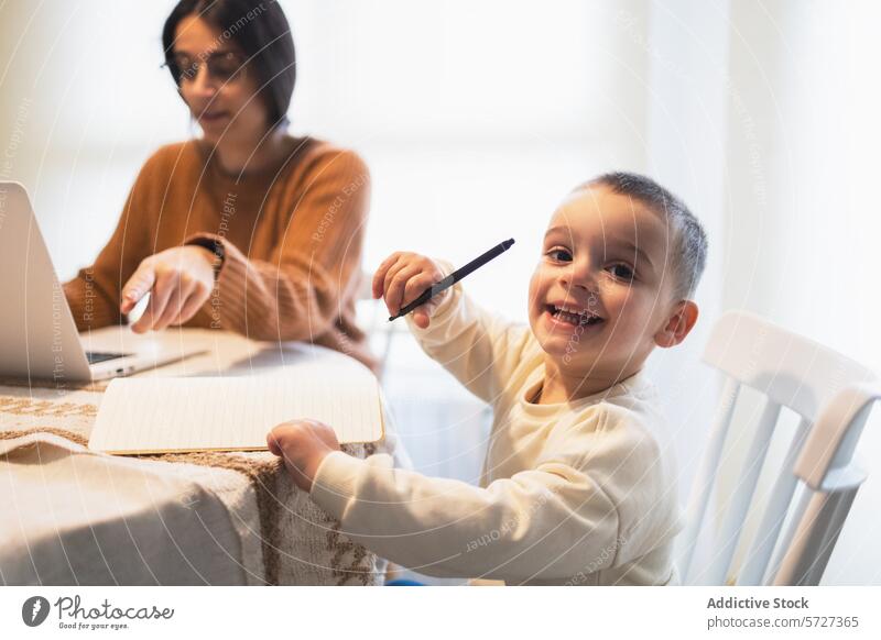 A cheerful young boy grins at the camera, holding a pen while sitting at a table with his mother, who is focused on her laptop toddler smiling work home family