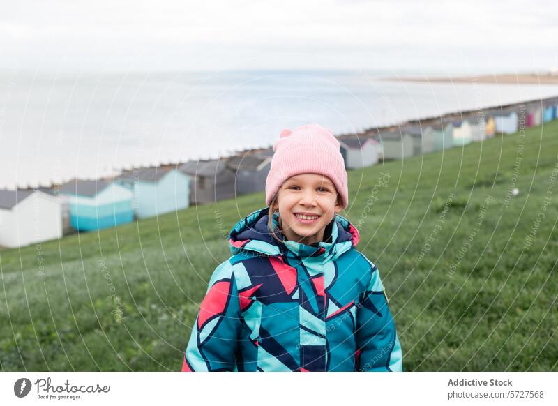 A cheerful young girl in a pink beanie and colorful coat smiles warmly on a green hill overlooking the iconic seaside huts of Whitstable, UK child portrait
