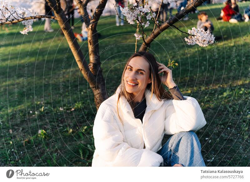 Smiling cheerful woman in white down jacket and jeans sitting on grass in city park in spring under cherry blossom young garden nature outdoor blooming beauty