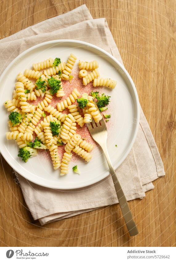 Elegant speckled plate holding a serving of fusilli pasta with broccoli florets, paired with a gold fork on a beige napkin and wooden background meal Italian
