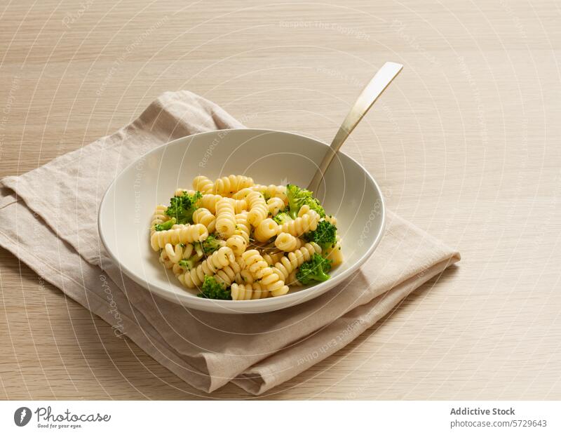 A serene setting with a bowl of fusilli pasta topped with steamed broccoli, accompanied by a beige linen napkin and a modern fork on a wooden surface topping
