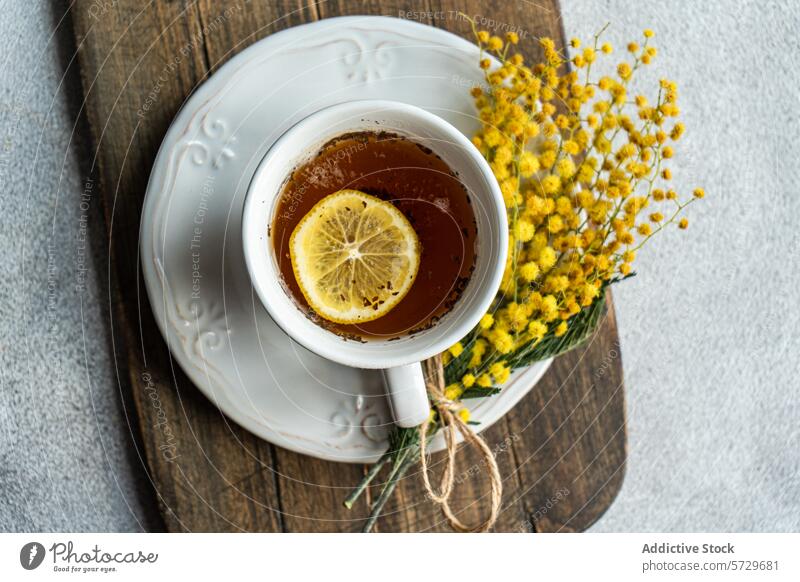 Top view of inviting cup of tea with a bright lemon slice on an ornate saucer, accompanied by a delicate Mimosa flower bundle, presented on a wooden board with a textured backdrop