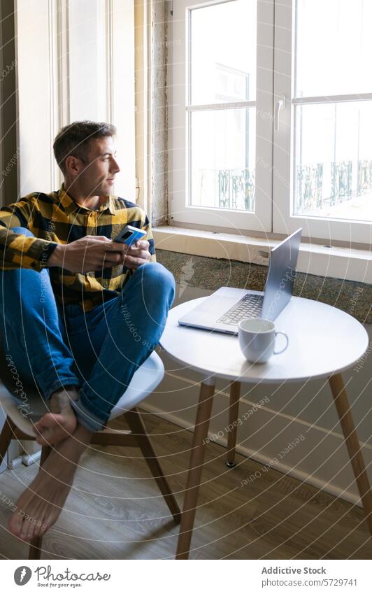 Man enjoying coffee break while working at home man barefoot window sipping smartphone laptop table indoor relaxation technology remote work telecommuting