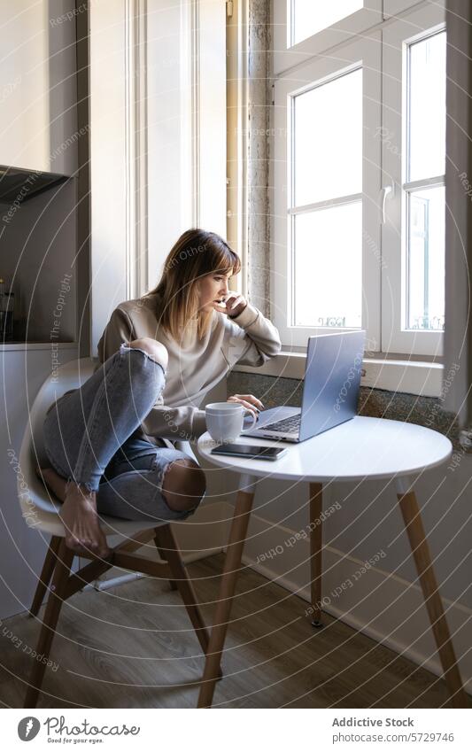 Young woman working on laptop at home with coffee table young seated window focus computer cup round table white table home office telecommute remote work