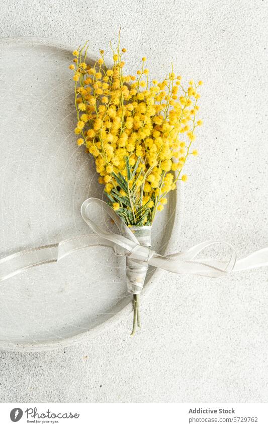 Top view of delicate arrangement of vibrant mimosa flowers tied with a silver ribbon, presented on a sleek, textured table surface for a sophisticated touch
