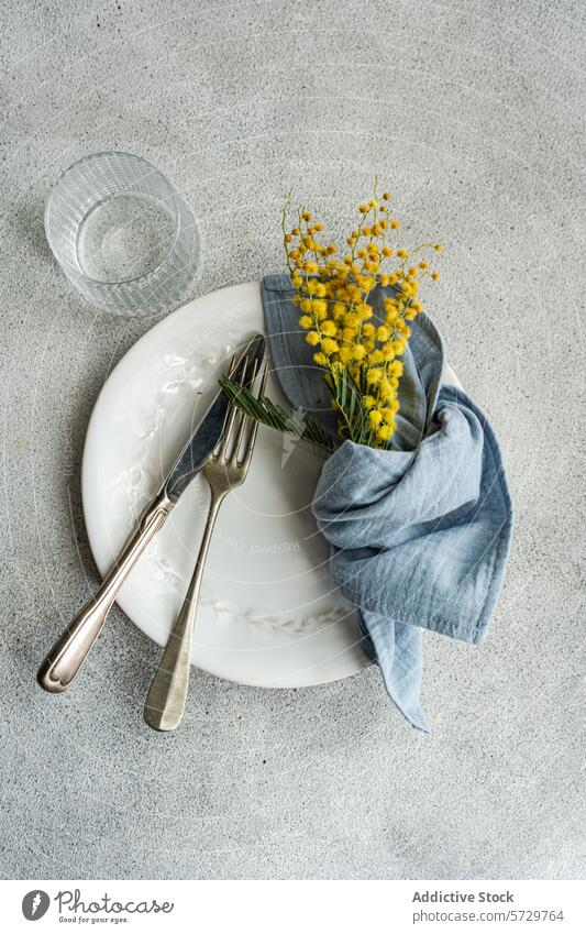 Elegant dining setup featuring a white ornate plate, vintage silverware, a blue linen napkin, and a bright yellow mimosa bouquet on a textured backdrop table