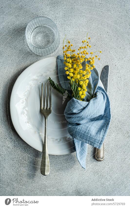 Top view of fresh outdoor table setting featuring a white embossed plate, silver cutlery, a blue linen napkin, and a sprig of bright yellow mimosa flowers decor