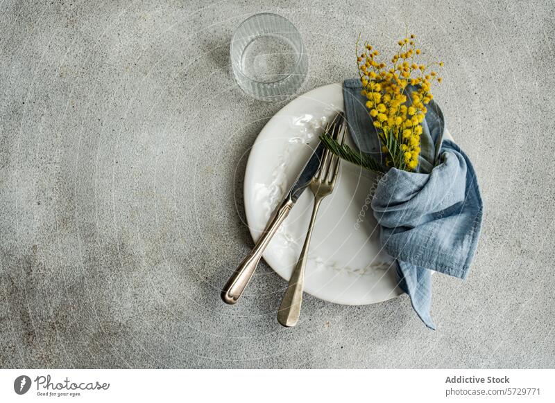 Elegant dining setup featuring a white ornate plate, vintage silverware, a blue linen napkin, and a bright yellow mimosa bouquet on a textured backdrop table