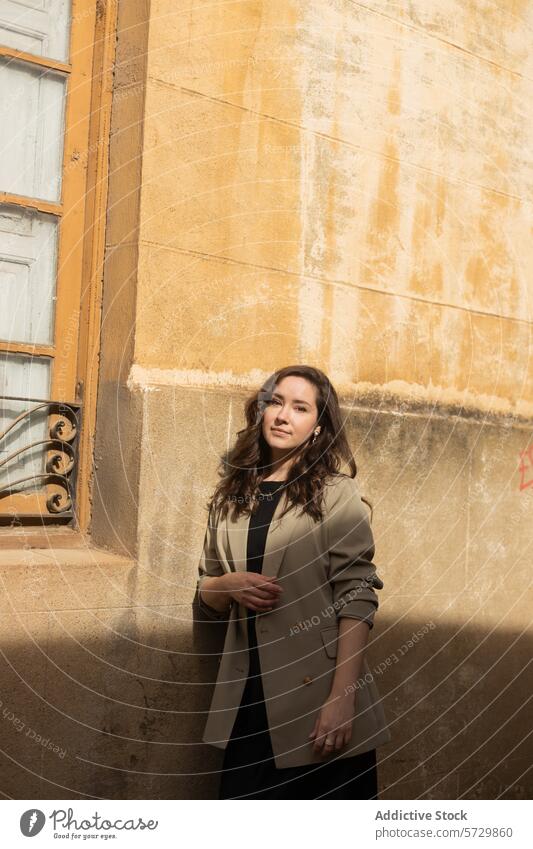A confident woman in a fashionable coat poses against a weathered wall with rustic charm and a hint of urban decay chic city texture style elegance casual