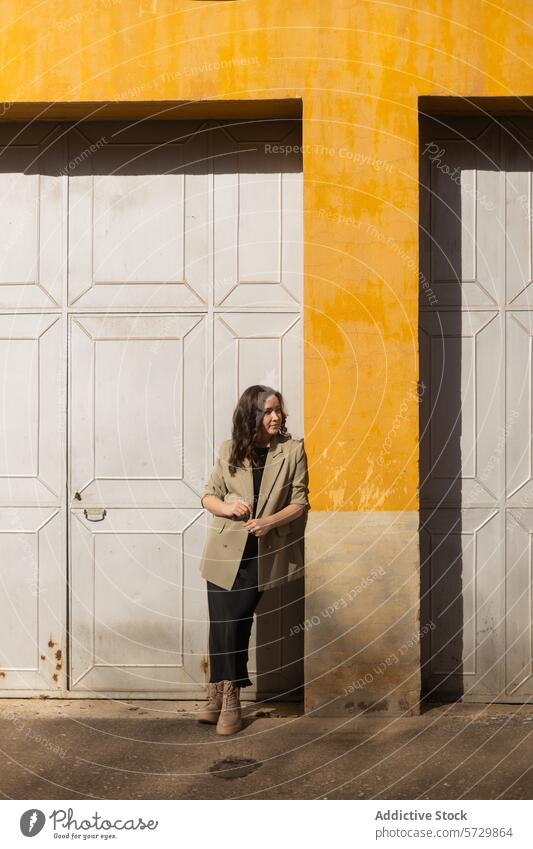 A thoughtful woman in a stylish trench coat stands beside vibrant yellow columns, basking in soft sunlight standing shadow architectural texture outdoor urban
