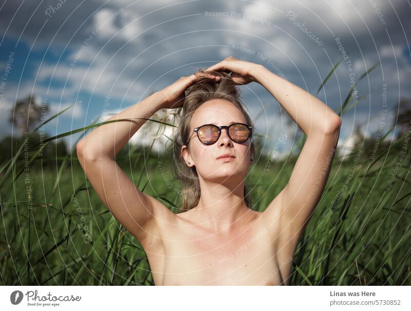 A gorgeous topless girl with sunglasses is having a relaxed afternoon surrounded by green nature and some dark clouds. A sexy image of a pretty woman who is feeling comfortable in her skin. Blurred lines might be a proper name for it.