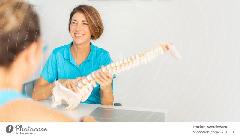 A physiotherapist explains to her patient the problem of low back pain and impingement in the vertebrae using an anatomical skeleton clinic rehabilitation