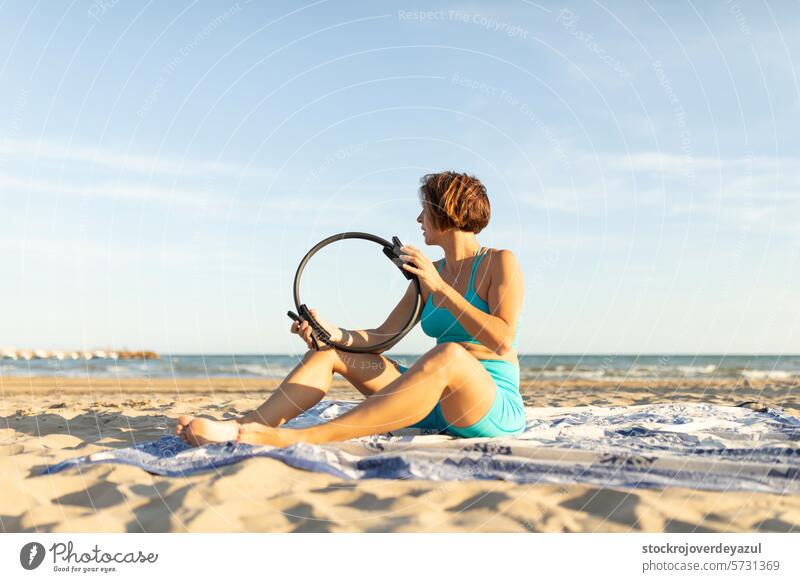 A woman rests sitting on the beach, during a series of exercises with the Pilates ring pilates yoga mediterranean spain mind-body exercise contrology sunset