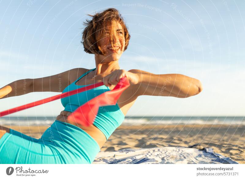 A woman smiles happily while doing Pilates exercises on the beach, in the sun and outdoors, with an elastic band pilates yoga mediterranean spain